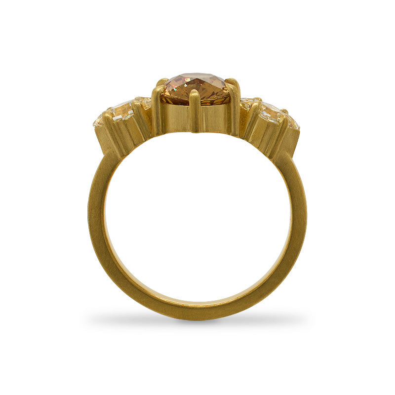 
                  
                    Load image into Gallery viewer, Through the finger view of an oval cut, orange sapphire ring with 2 lozenge cut diamonds set on either side of the sapphire, and set in a claw prong 18 kt yellow gold setting with a matte finish on a white background.
                  
                