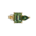 Front view of an asymmetrical, emerald cut, green tourmaline ring flanked by one half moon cut white sapphire, one pear cut green sapphire, two round cut white diamonds, and cast in 14 kt yellow gold.