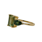 Right side view of an asymmetrical, emerald cut, green tourmaline ring flanked by one half moon cut white sapphire, one pear cut green sapphire, two round cut white diamonds, and cast in 14 kt yellow gold.