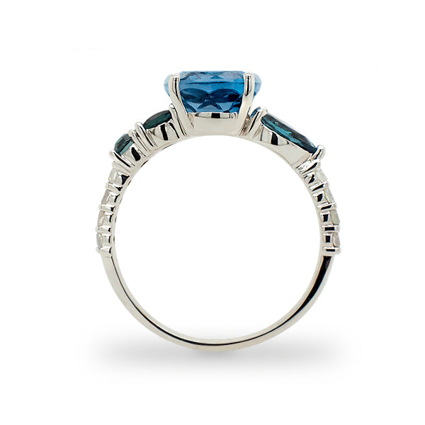Side view of cushion cut, London blue topaz with 2 round and 1 pear cut green tourmaline stones, and 8 round cut diamonds going down the band cast in 14 kt white gold.