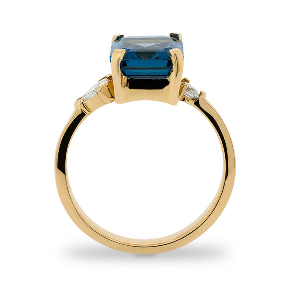 Side view of emerald cut London blue topaz and round and trillion cut diamond ring cast in 14 kt yellow gold.