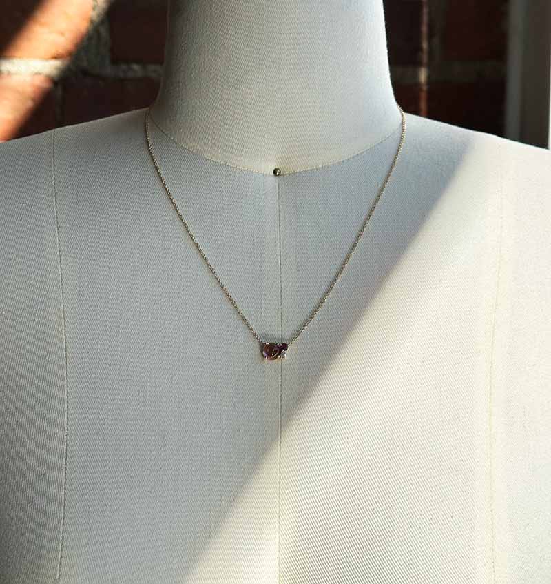 Front view of a cabochon cut, pink tourmaline necklace with one round cut, faceted, deep red tourmaline and one round cut diamond necklace cast in 14 kt yellow gold on a body form for scale.