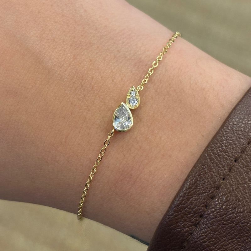 A yellow gold bezel pear crystal next to two small round crystals in a smaller pear setting along a sparkly cable chain. Shown on a model's right wrist.