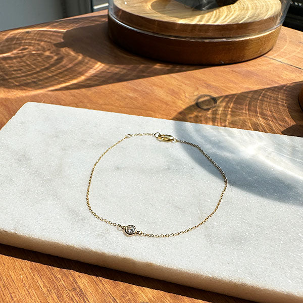 
                  
                    Load image into Gallery viewer, Overview shot of a solitaire, bezel set diamond bracelet made of solid 14 kt yellow gold on top of an ivory tile resting on a wooden tabletop.
                  
                