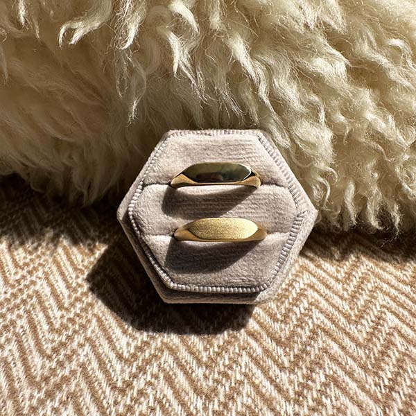 One 14 kt gold high polish and one matte finish signet ring in a ring holder with a woolen pillow and chevron style pattern blanket in the background.