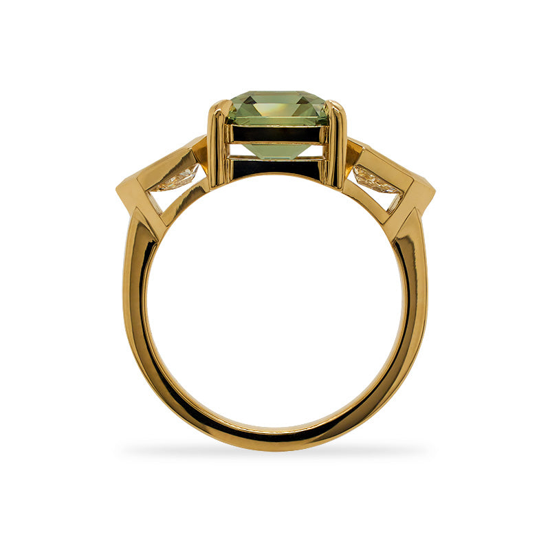 A "through the finger" view of a prong set, asscher cut, green sappihire and bezel set shield cut diamond ring in an 18 kt yellow gold setting.