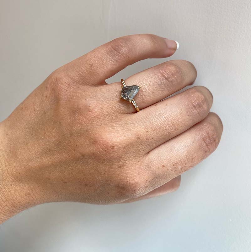 Front view on left ring finger of a 2.39 ct, kite cut, salt and pepper diamond in an 18 kt yellow gold claw prong setting with 3 pavé set diamonds on each side of the band.