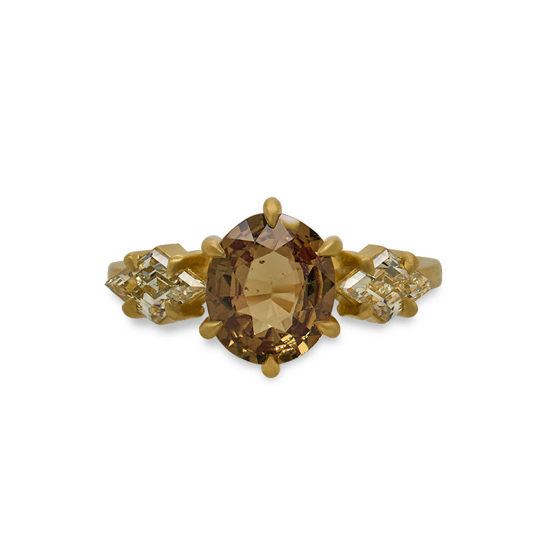 Front view of an oval cut, orange sapphire ring with 2 lozenge cut diamonds set on either side of the sapphire, and set in a claw prong 18 kt yellow gold setting with a matte finish on a white background. 