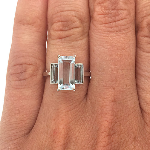 A three stone aquamarine ring with one larger, emerald cut center stone flanked by two baguette cut stones set in a 14 kt white gold setting on wearer's left ring finger for scale.