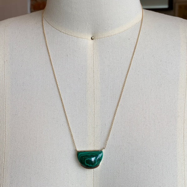 Front view of malachite necklace on dress form suspended on 14 kt yellow gold chain on dress form. Malachite is cut in half moon shape and is partially bezel set on top and bottom of stone. 