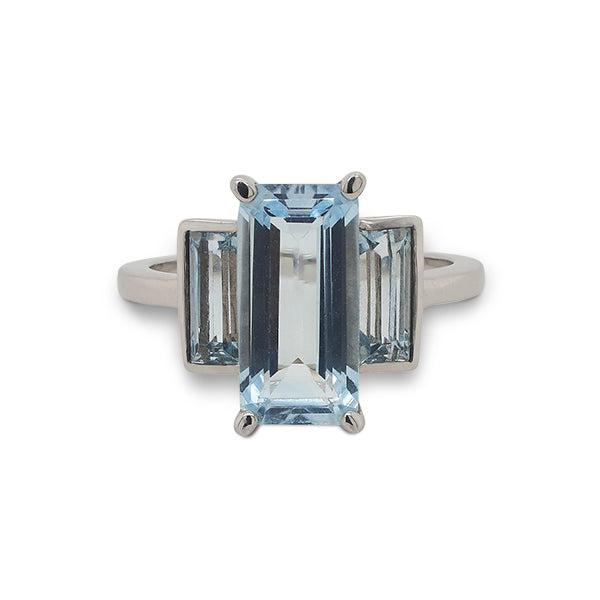 Front view of a three stone aquamarine ring with one larger, emerald cut center stone flanked by two baguette cut stones set in a 14 kt white gold setting.
