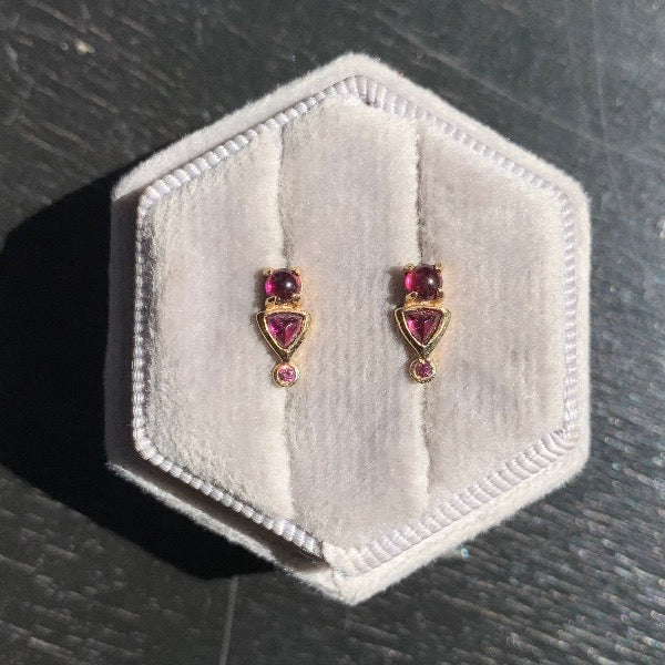 Front view on grey velvet box of rhodolite garnet stud earrings with round, cabochon, and trillion cut stones set in 14 kt yellow gold settings. Photo in natural sun light.