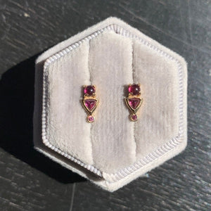 
                  
                    Load image into Gallery viewer, Front view on grey velvet box of rhodolite garnet stud earrings with round, cabochon, and trillion cut stones set in 14 kt yellow gold settings. Photo in natural sun light. Displayed on a light gray velvet earring box on a dark gray table.
                  
                