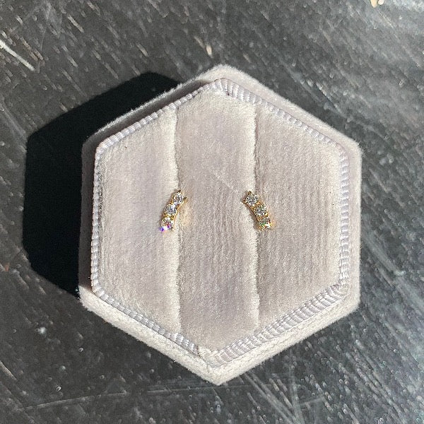 Front view of round diamond studs,  0.18 TCW, each stud prong set 3 in a row in a slight curve. Cast in 14 kt yellow gold. Photo taken on grey velvet in natural sunlight.