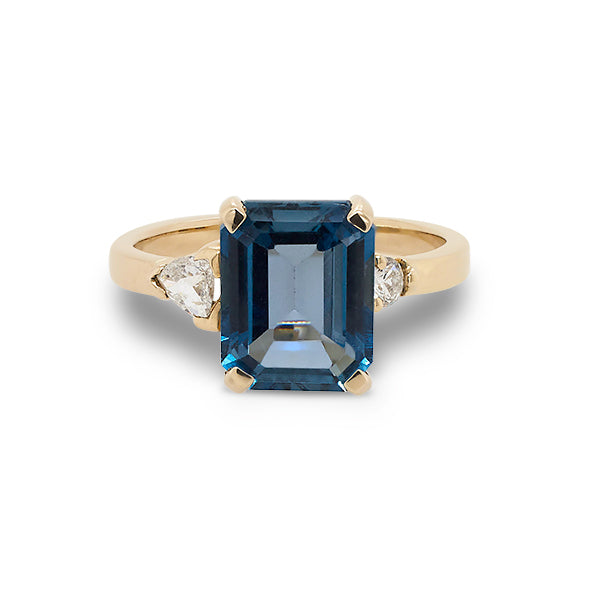 Front view of emerald cut London blue topaz and round and trillion cut diamond ring cast in 14 kt yellow gold.