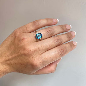
                  
                    Load image into Gallery viewer, Front view on left ring finger of emerald cut London blue topaz and round and trillion cut diamond ring cast in 14 kt yellow gold.
                  
                
