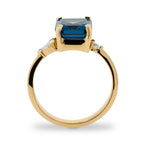 Side view of emerald cut London blue topaz and round and trillion cut diamond ring cast in 14 kt yellow gold.