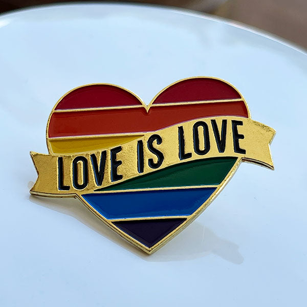 Front view of enamel heart shaped pin with rainbow stripe and gold toned banner waving across center with 