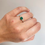Front view on left ring finger of round emerald ring with 2 medium and 6 small accent diamonds cast in 14 kt yellow gold.