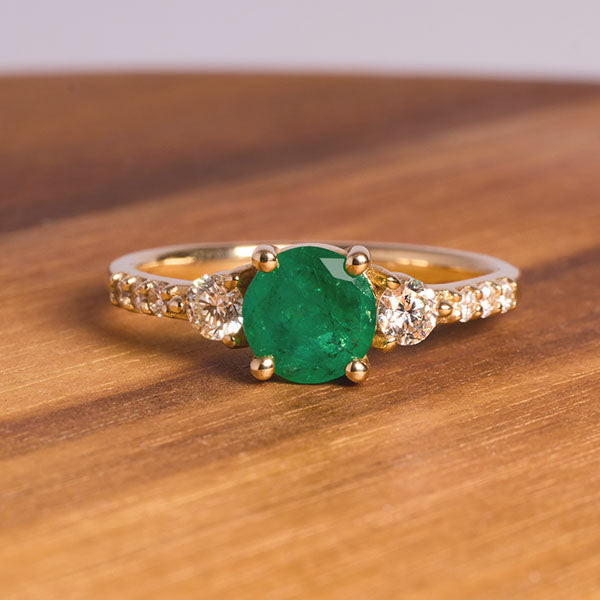 
                  
                    Load image into Gallery viewer, Front view of round emerald ring with 2 medium and 6 small accent diamonds cast in 14 kt yellow gold on wooden desktop.
                  
                