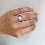 Front view on left ring finger of an asymmetrical ring with a large cabochon moonstone flanked by a smaller moonstone, diamond and half moon cut blue topaz and cast in 14 kt yellow gold.