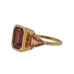 This right side view of a ring is cast in 18 kt yellow gold has a large 3.5 carat emerald cut rhodolite garnet in the center and is flanked by almost 2 carats of peach colored emerald and trillion cut tourmalines and tiny round cut diamonds.
