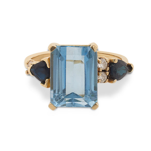 Front view of an asymmetrical cut light blue topaz, pear cut sapphire and double diamond ring cast in 14 kt yellow gold.