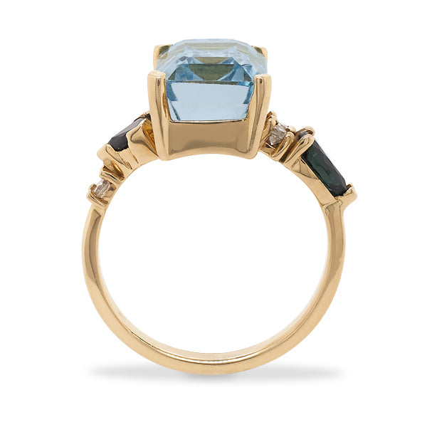 Side view of an asymmetrical cut light blue topaz, pear cut sapphire and double diamond ring cast in 14 kt yellow gold.