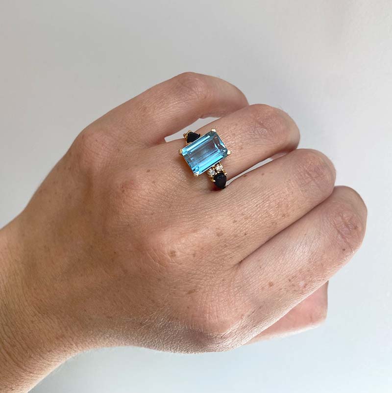 
                  
                    Load image into Gallery viewer, Front view on left ring finger of an asymmetrical cut light blue topaz, pear cut sapphire and double diamond ring cast in 14 kt yellow gold.
                  
                