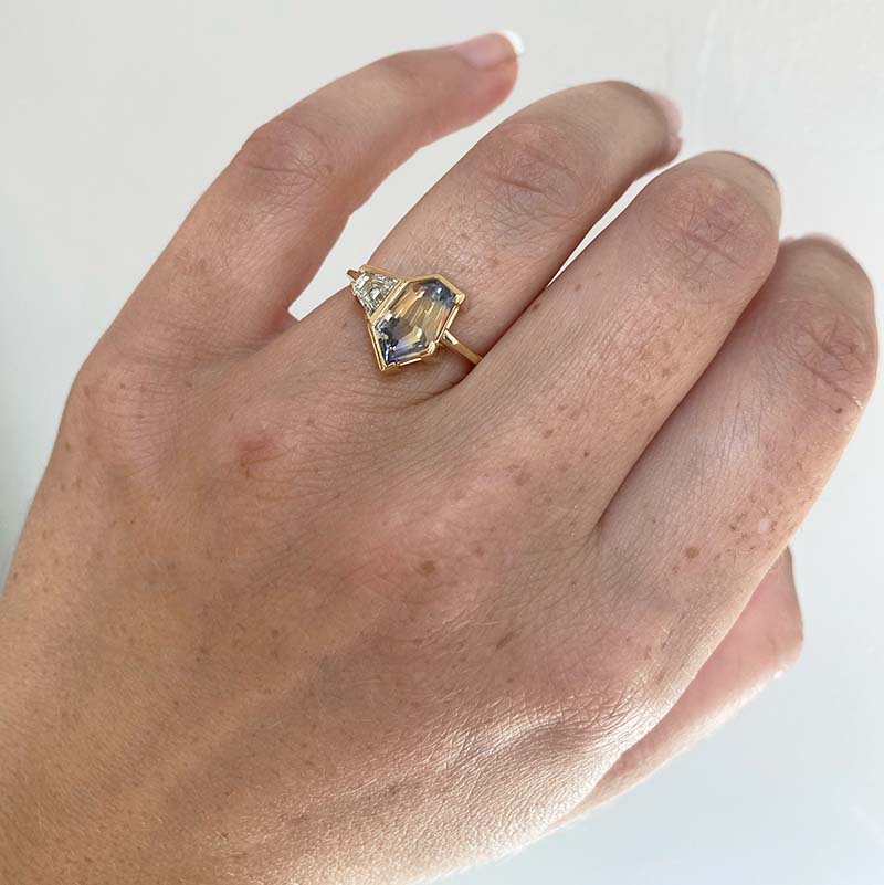 Front view on left ring finger of a light blue with yellow bicolor sapphire, cut in an irregular geometric shape with a trapezoid cut white diamond set alongside. Cast in 18 kt yellow gold.